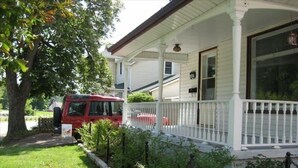 Une maison avec un porche blanc, un véhicule rouge garé devant, et un arbre sur la gauche.
