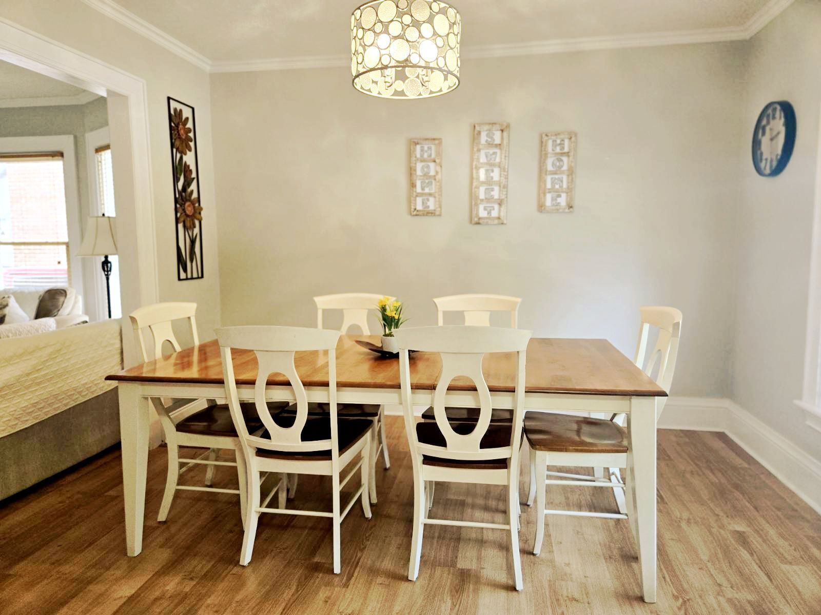 Un coin repas avec une table en bois et des chaises blanches, un luminaire suspendu et des tableaux encadrés au mur.