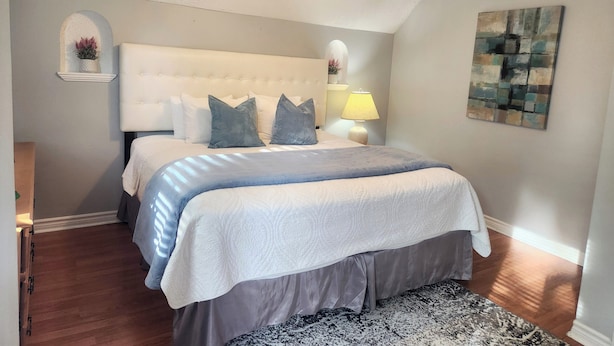Une chambre à coucher avec une tête de lit capitonnée, un lit recouvert d’une couette grise et blanche, un tapis à motifs, un ventilateur de plafond et une lampe fixée au mur.