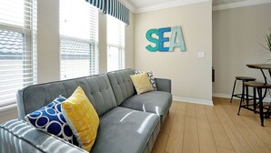 Uma sala de estar com sofá cinzento, almofadas estampadas, uma placa na parede com a palavra 