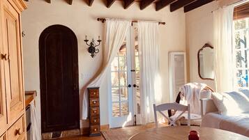 A cozy room with a wooden door, a white chair, a round wooden table, and a bed with white bedding.