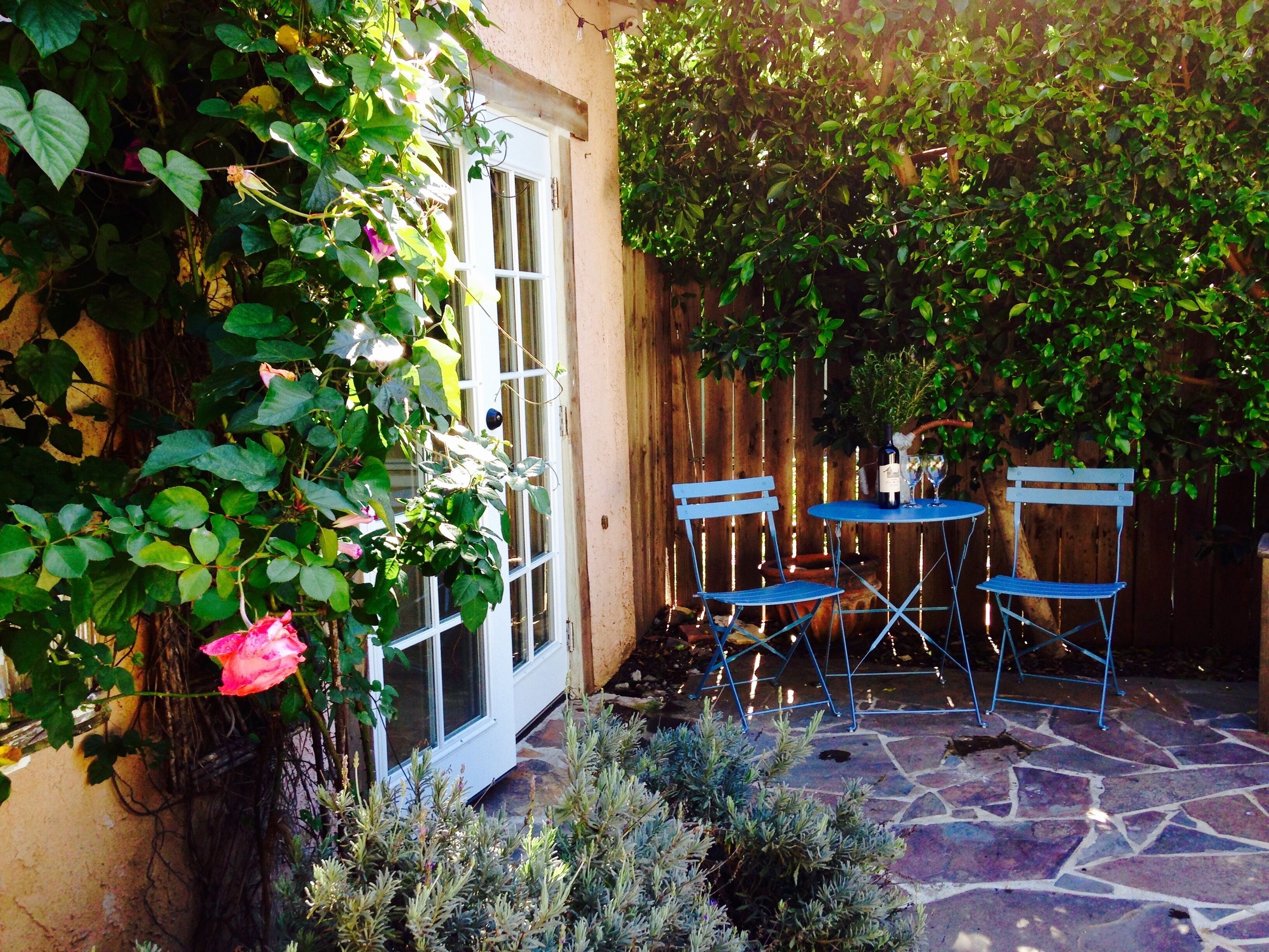 Une terrasse avec un sol en pierre, deux chaises en métal bleues et une table sur laquelle se trouvent une bouteille et des verres.