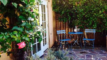 Une terrasse avec un sol en pierre, deux chaises en métal bleues et une table sur laquelle se trouvent une bouteille et des verres.