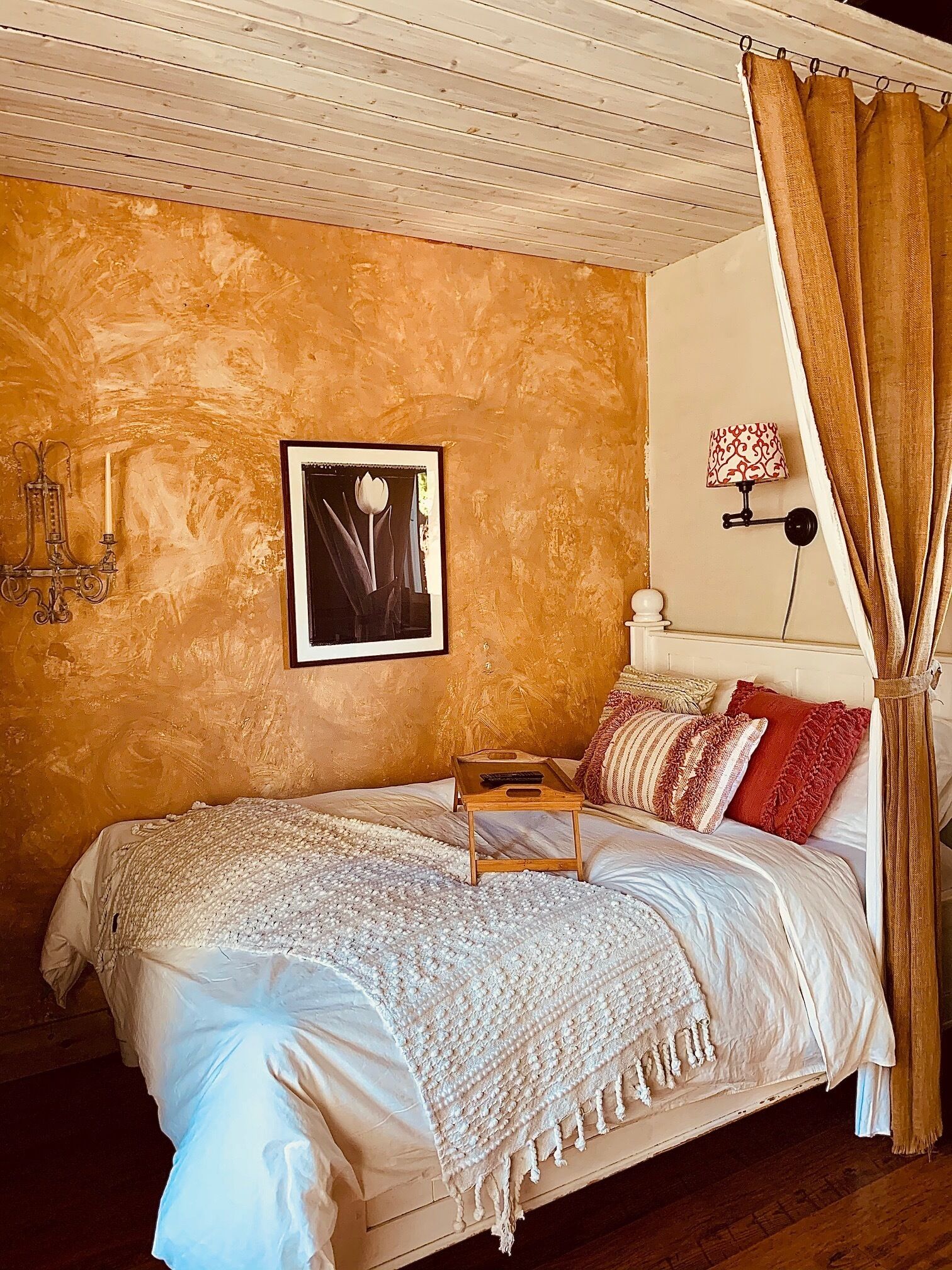 A bed with a wooden tray, a framed picture, a wall-mounted lamp, and a curtain.