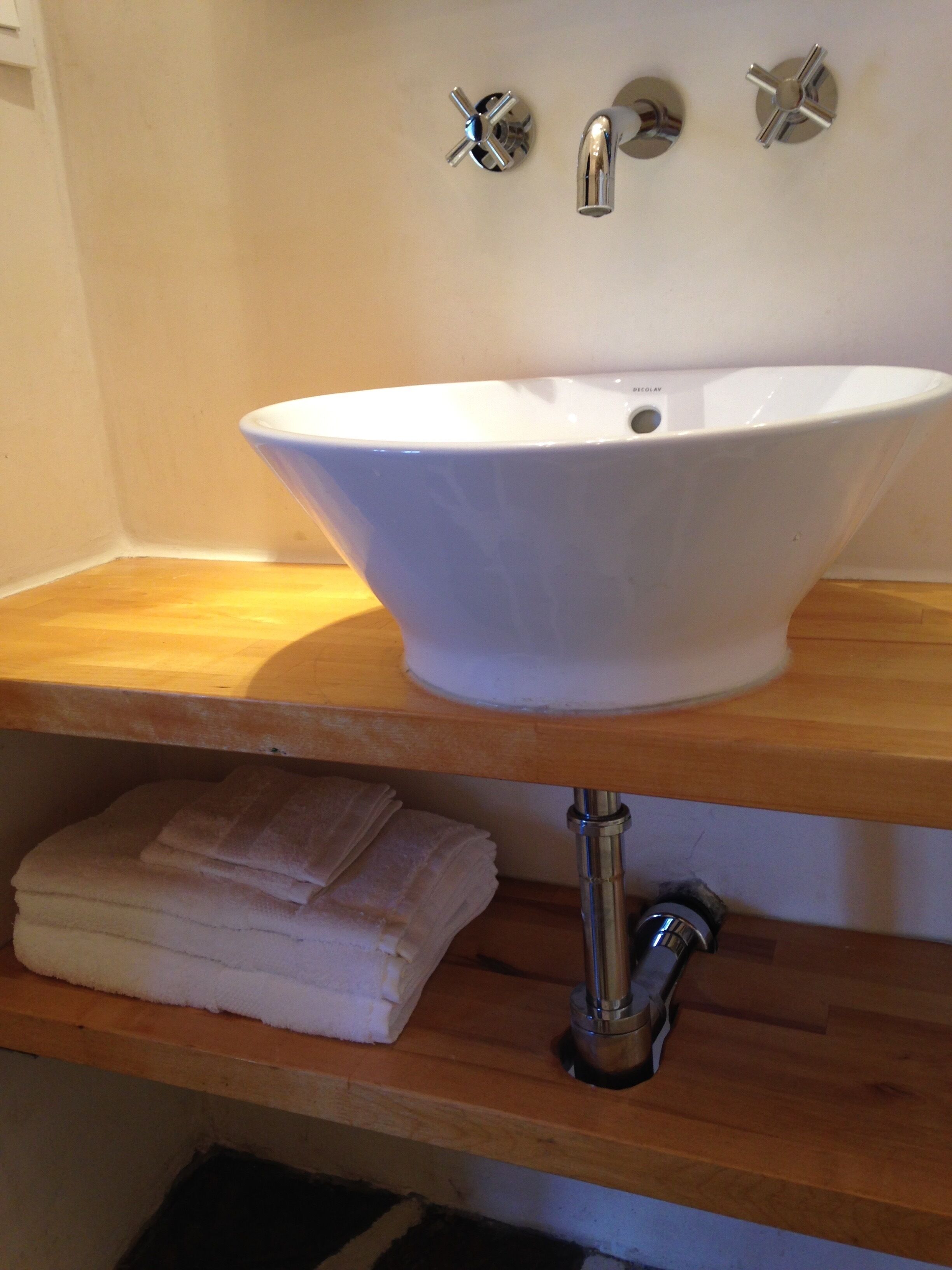 A bathroom with a white sink, two chrome faucets, and a wooden shelf with folded towels.