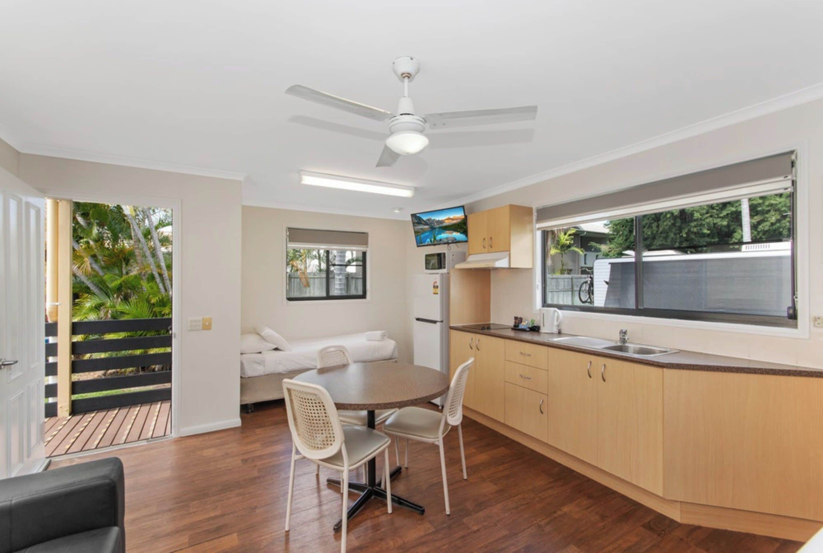 A modern kitchen with wooden cabinets, a dining area with a round table and chairs, a ceiling fan, and a large window.