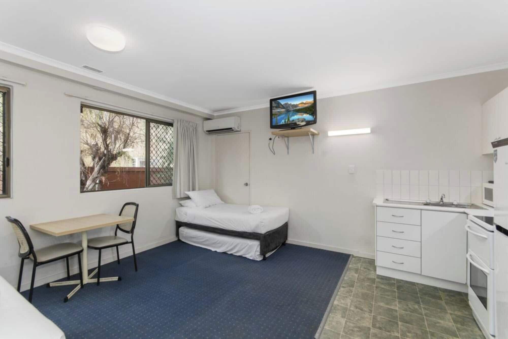 A compact room with a bed, a desk, a chair, a TV, and a kitchenette.