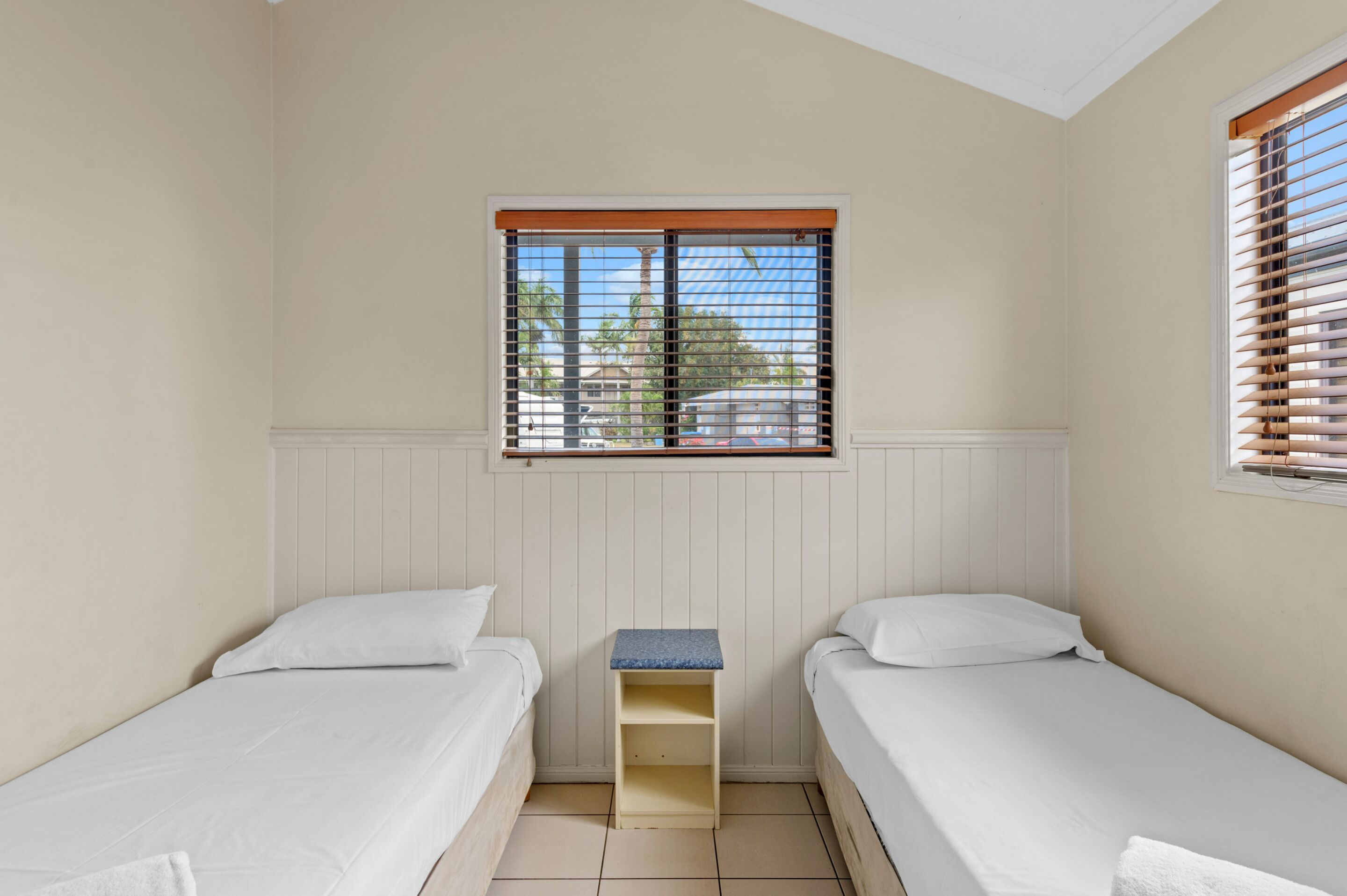 Een kleine, schone kamer met twee eenpersoonsbedden, een klein kastje en een raam met zonwering.