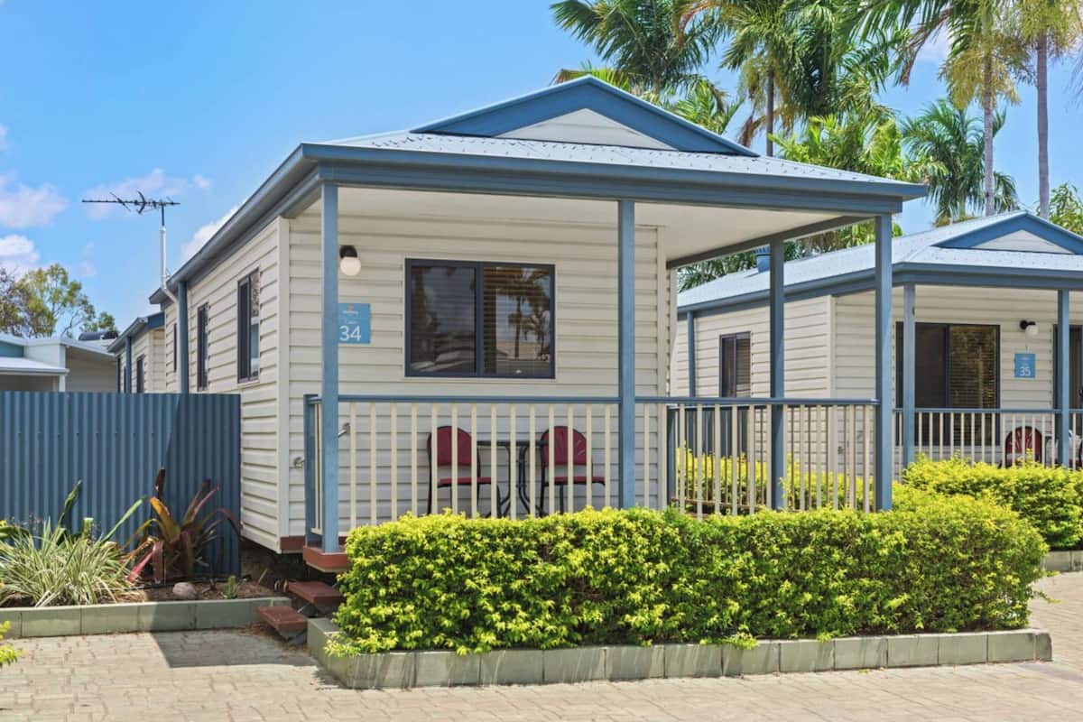 A single-story mobile home with a blue roof and beige siding, numbered 34. It features a covered porch with a railing and a small garden area with green shrubs.