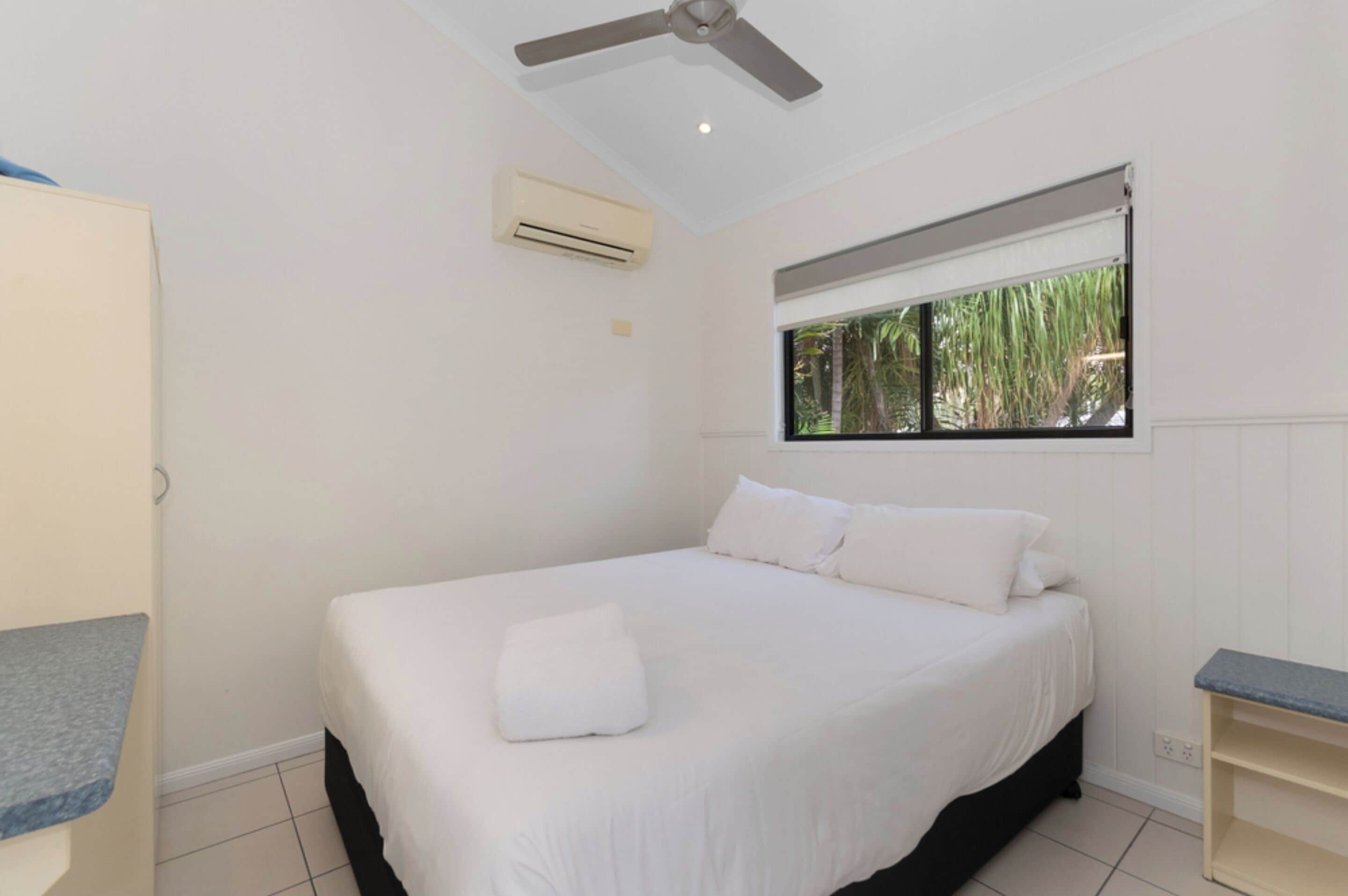 A bedroom with a bed, a ceiling fan, an air conditioning unit, a window with a view of greenery, and a small shelf.