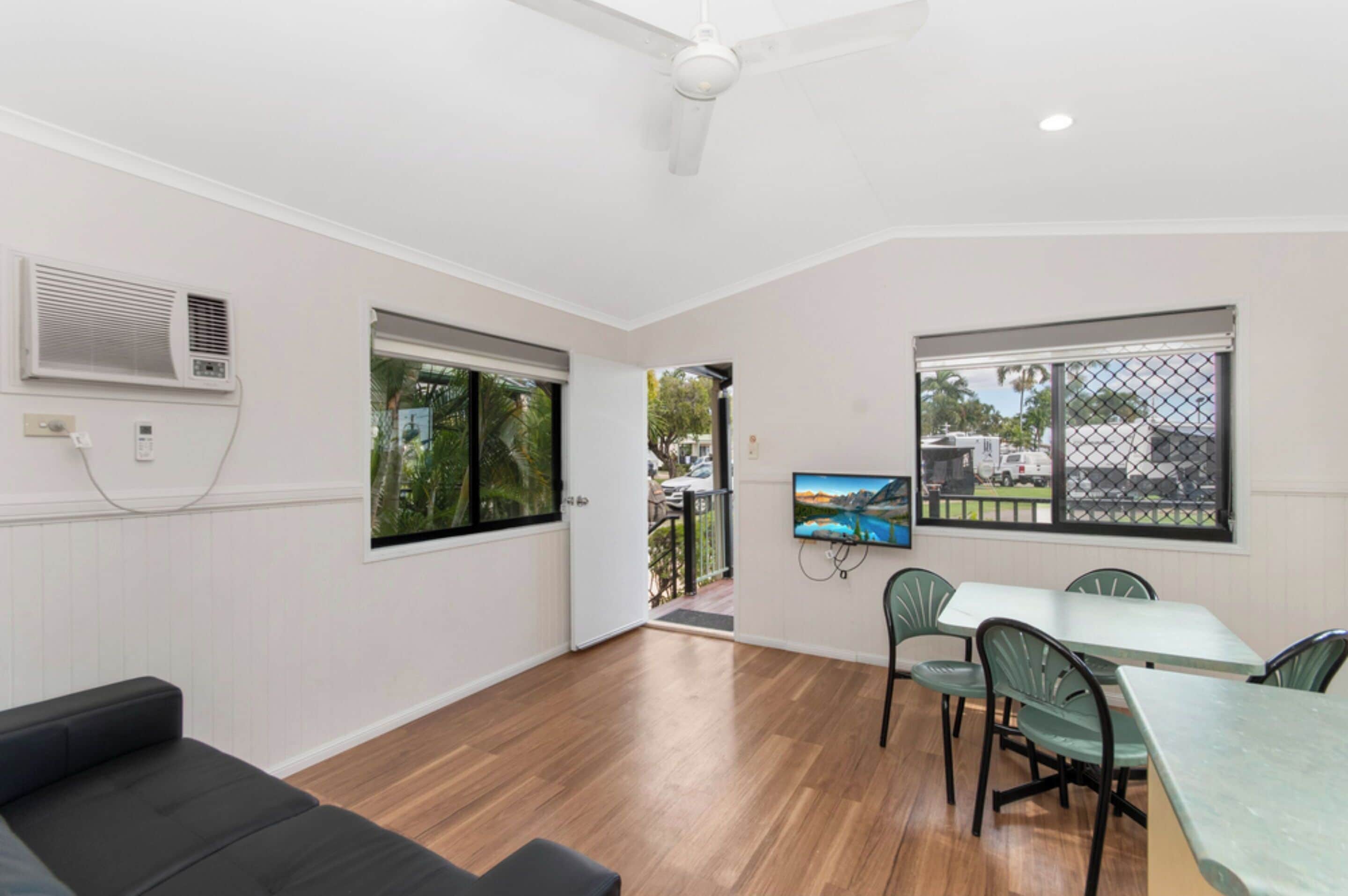 A modern room with a wooden floor, a ceiling fan, a large window with a view, a flat-screen TV, and a dining area with green chairs.