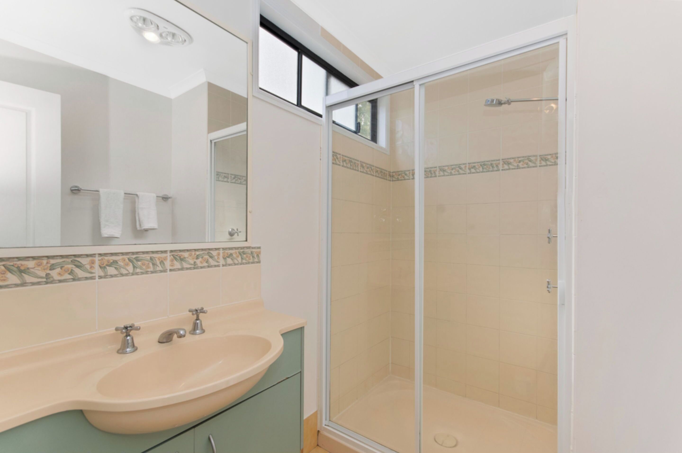 A bathroom with a beige sink, two faucets, a mirror, and a glass-enclosed shower.