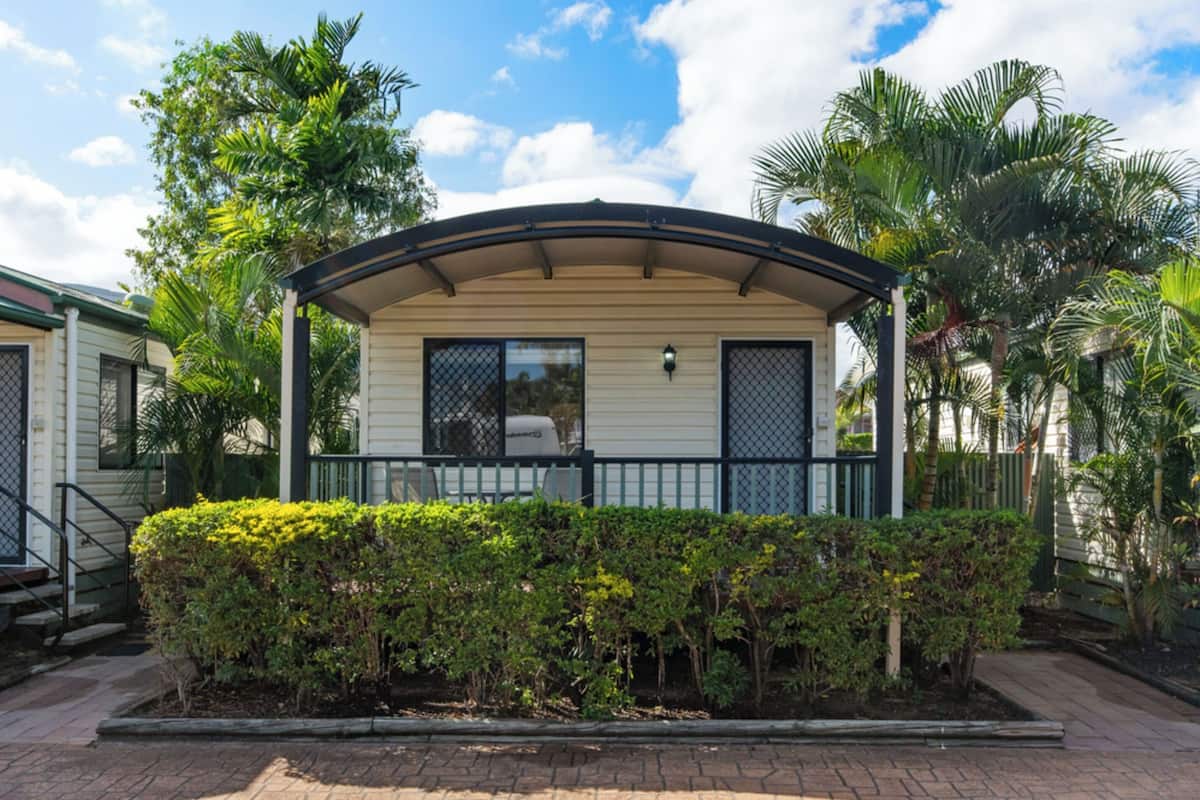 A single-story mobile home with a covered porch, surrounded by lush greenery and palm trees.