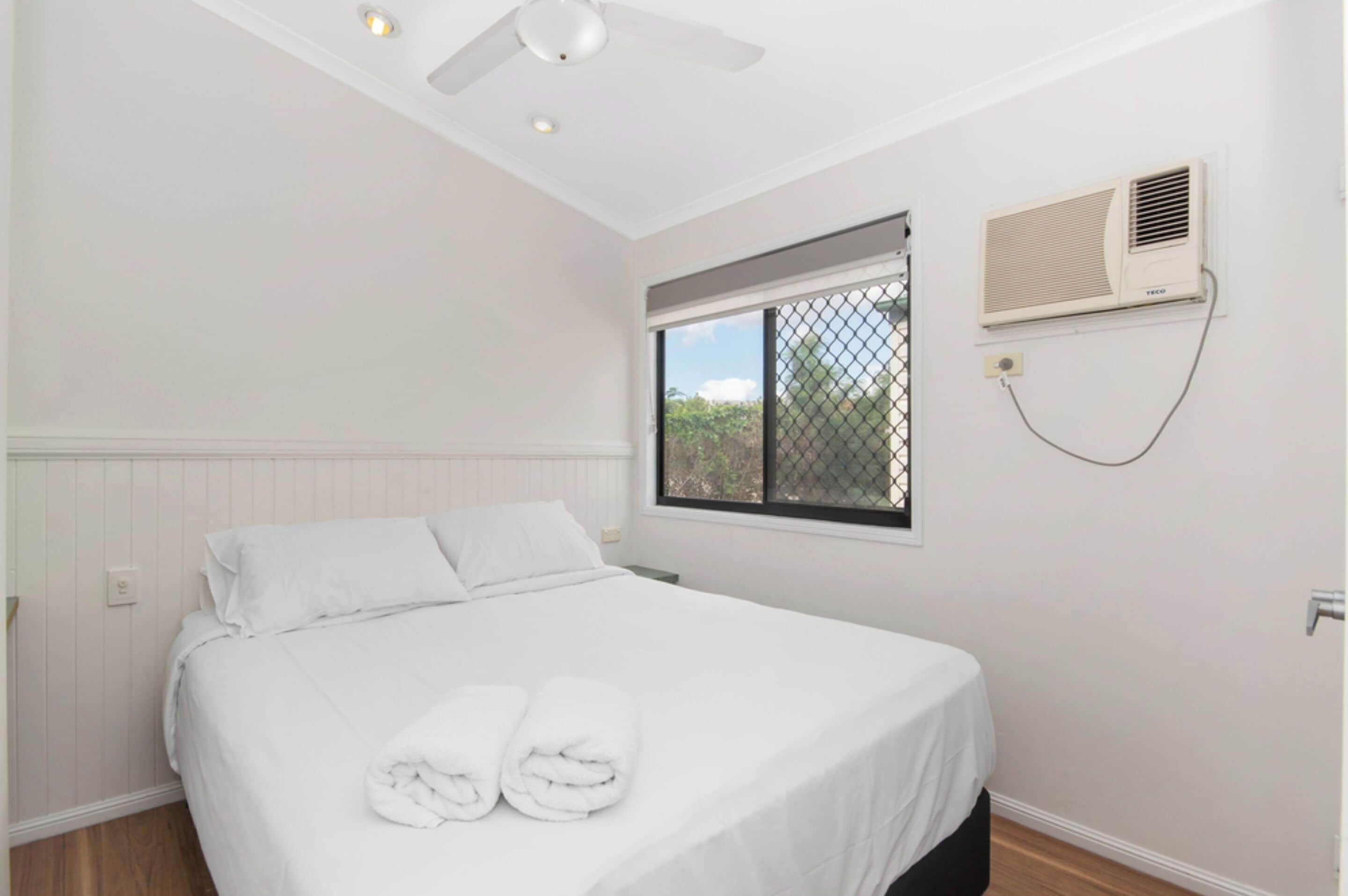 A bedroom with a bed, white linens, a ceiling fan, a window with a view, and an air conditioning unit.