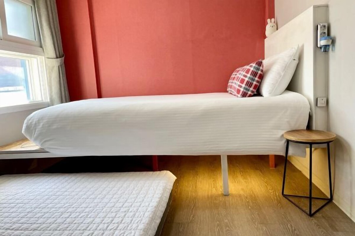 A single bed with a white mattress and a red and white pillow, a small round stool, and a window with curtains.