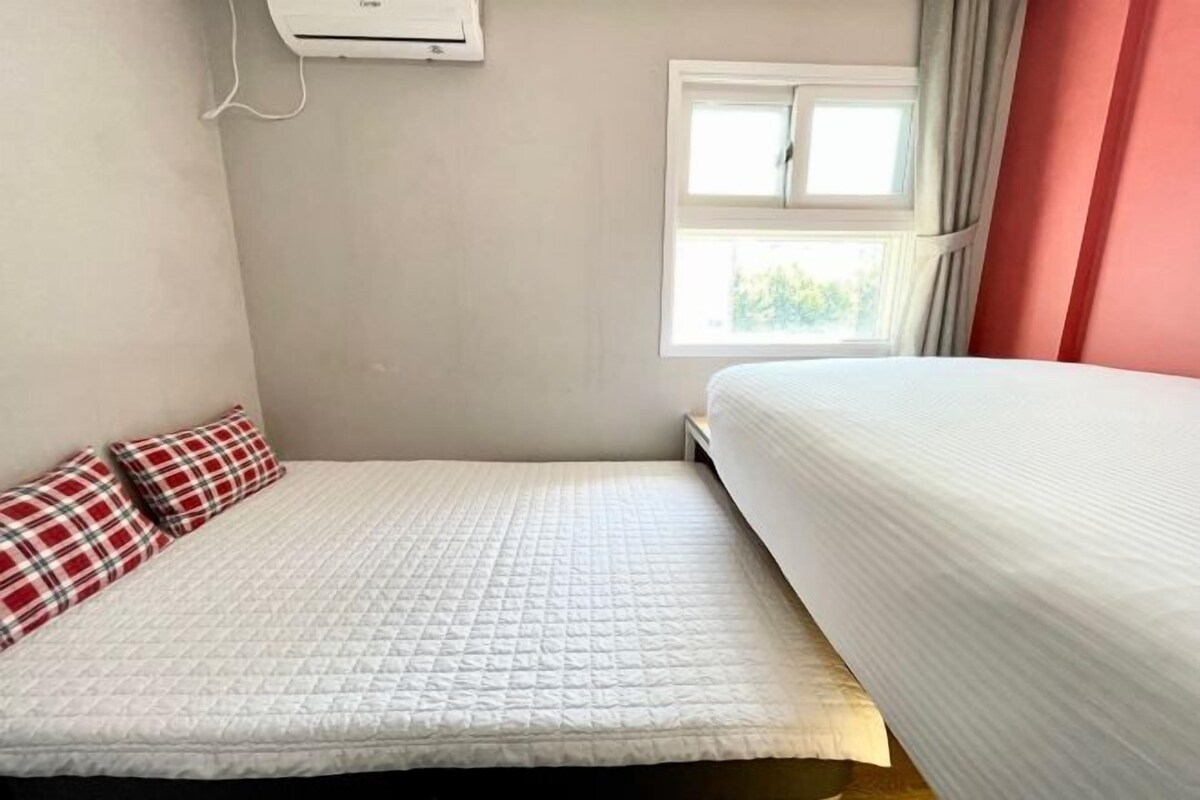 A small, single-bed room with a white quilted bedspread, a red and white plaid pillow, and a window with curtains.