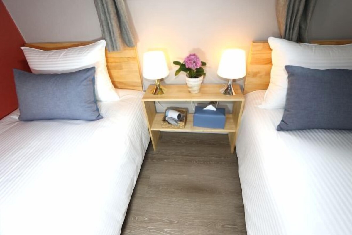 A hotel room with two single beds, each with a pillow and a bedside table with a lamp, a vase with flowers, and a small box.