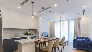 A modern kitchen with a central island, dining area with chairs, and a blue sofa in a living room with large windows. - Vinhomes Central Park - MM Home (Ho Chi Minh City)
