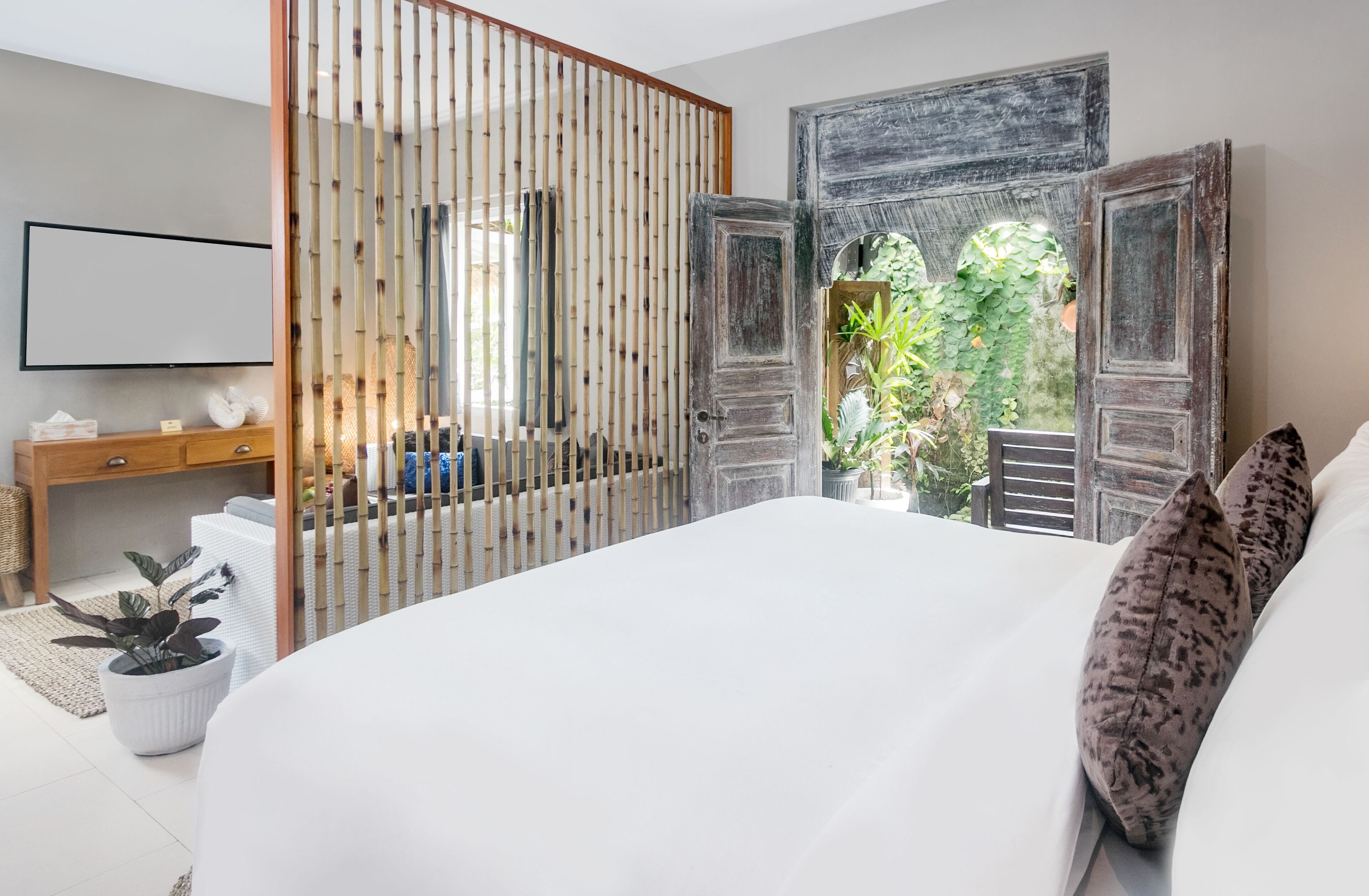A bedroom with a large bed, a wooden desk, and a view of greenery through open doors.