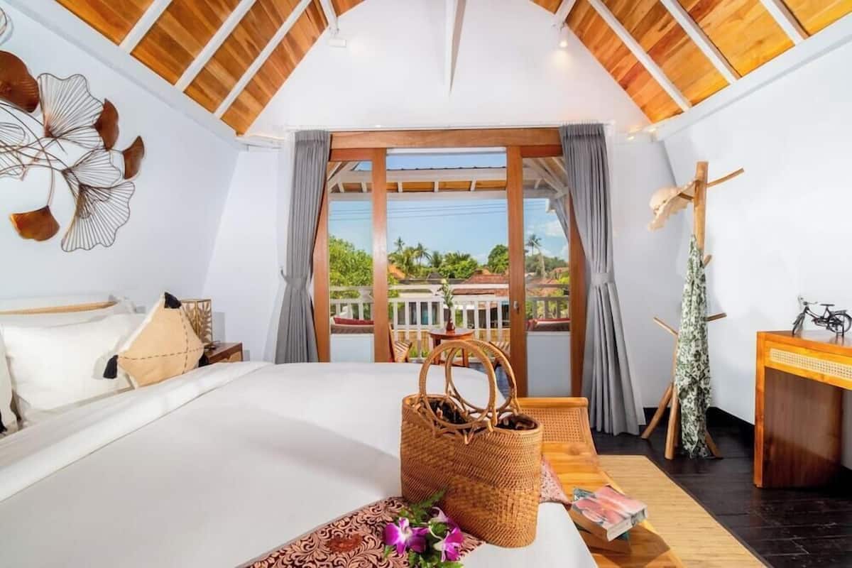 Un dormitorio con una cama grande, techo de madera, vista a través de un ventanal, una cesta de mimbre y un escritorio con una bicicleta.