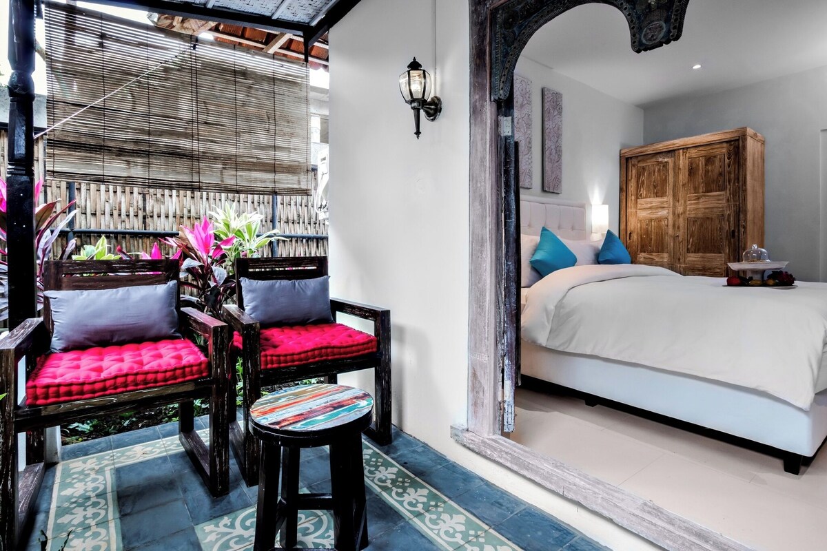 A room with a bed, a wooden wardrobe, a wall-mounted lamp, and a seating area with chairs and a small table.
