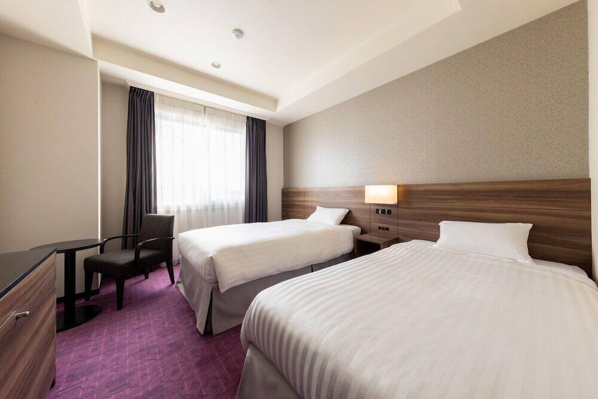 A hotel room with two beds, a desk, a chair, a window with curtains, and a headboard with a light.