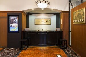 Una zona de recepción con mostrador de madera, dos taburetes tipo bar, una pantalla digital mostrando información, una obra de arte abstracta enmarcada y un reloj con mapa del mundo en la pared.
