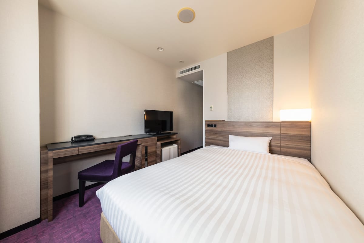 A hotel room with a bed, a desk with a chair, a television, and a headboard with a light.