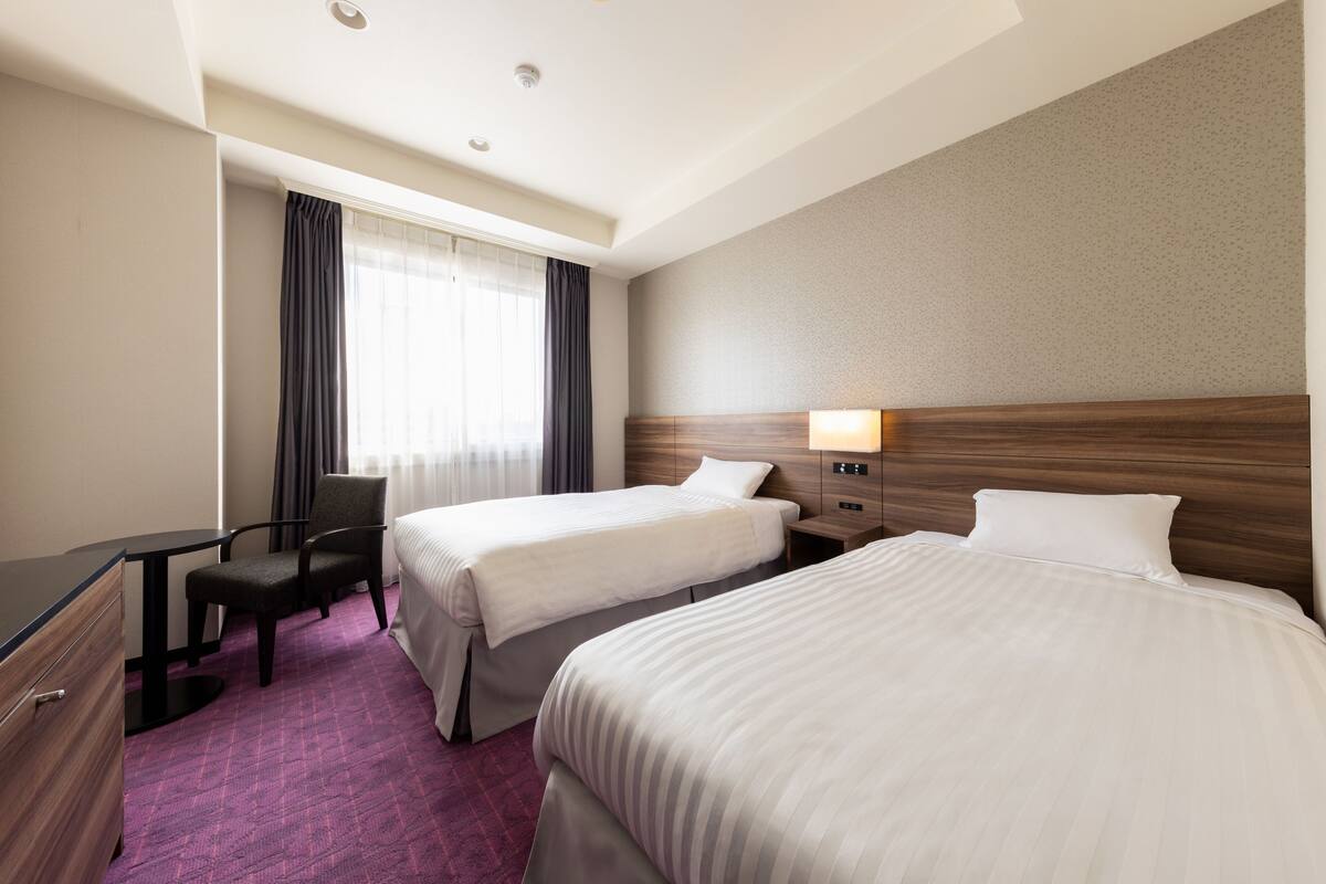 A hotel room with two beds, a desk, a chair, and a window with curtains.