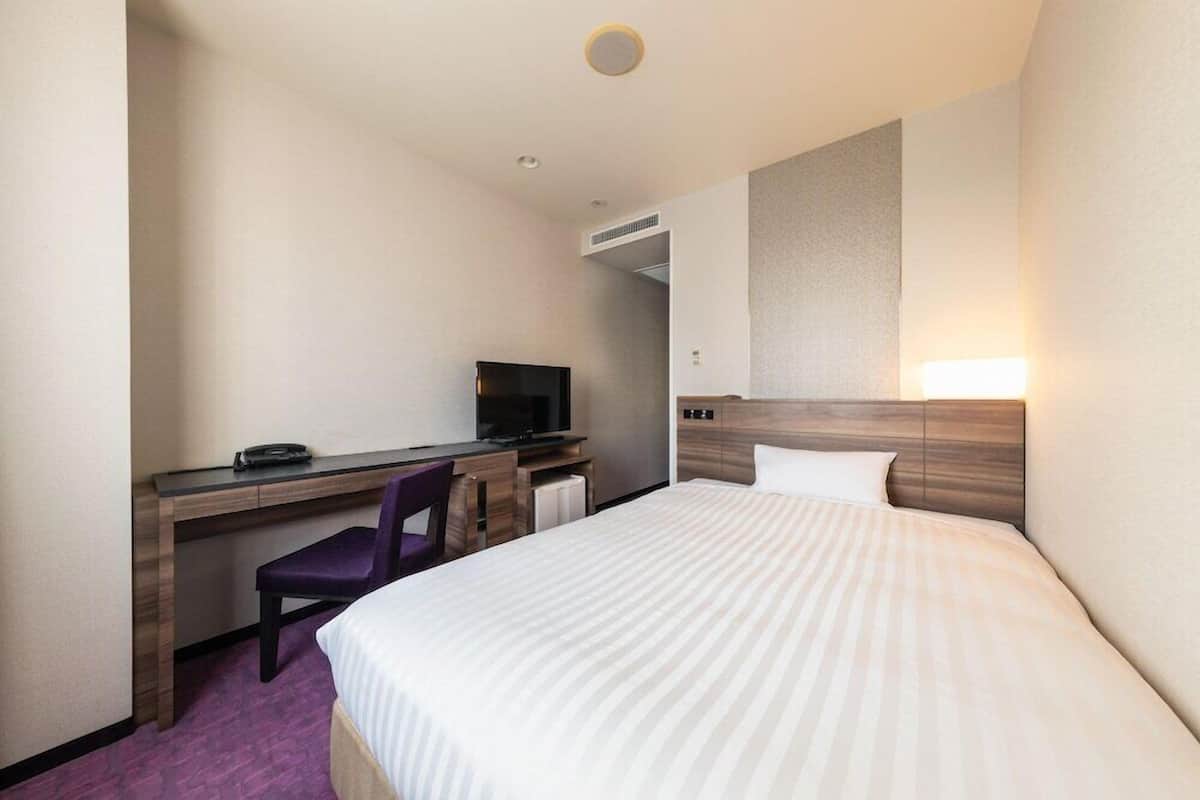 A hotel room with a bed, a desk with a chair, a television, and a wall-mounted light.