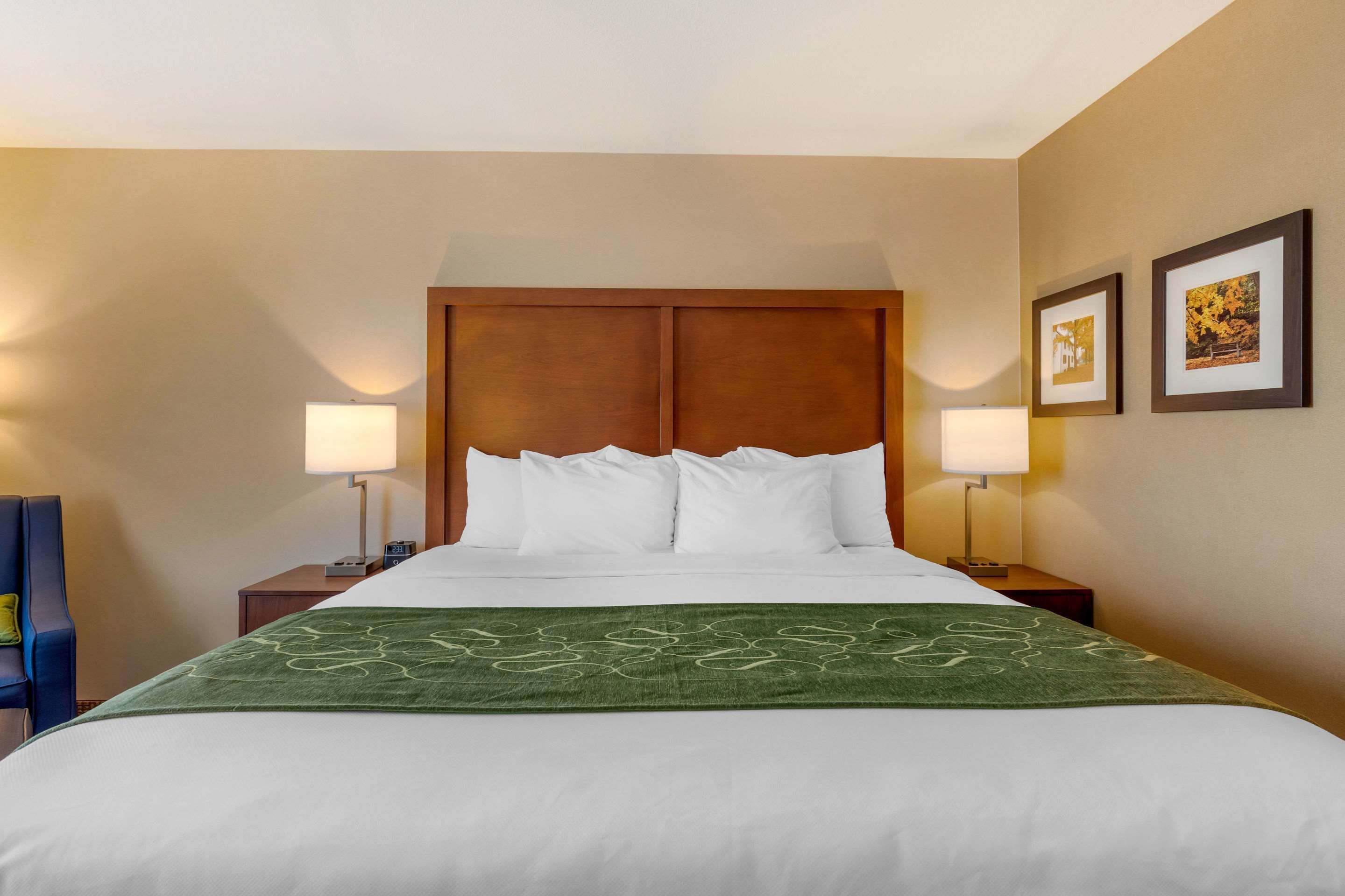 A hotel room with a large bed, two bedside tables with lamps, a blue chair, and two framed pictures on the wall.