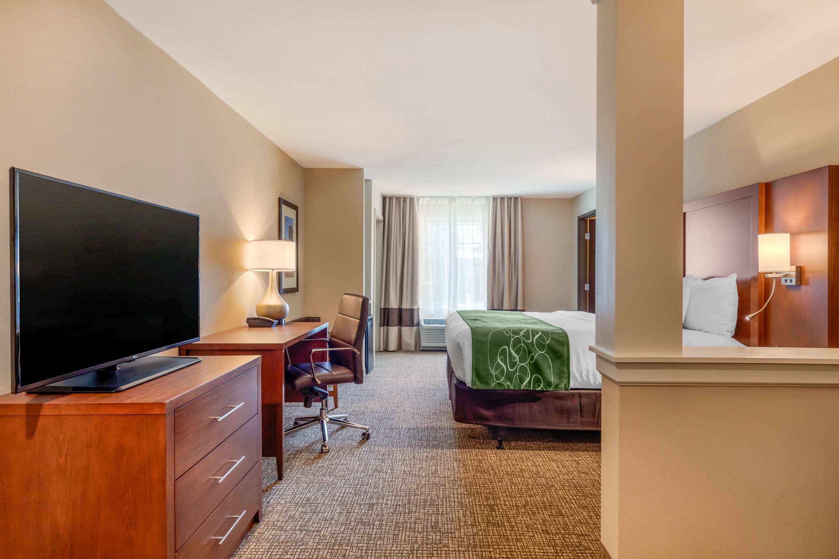 A hotel room with a flat-screen TV, a desk with a lamp, a chair, a bed with a green blanket, and a large mirror.