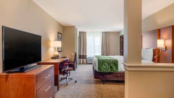 A hotel room with a flat-screen TV, a desk with a lamp, a chair, a bed with a green blanket, and a large mirror.