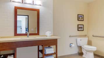 Un baño con lavamanos, espejo y inodoro blancos. Hay toallas en un estante y un cuadro colgado en la pared.