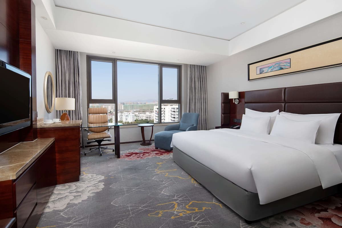 A hotel room with a large bed, a desk with a TV, a chair, and a view of the city through large windows.