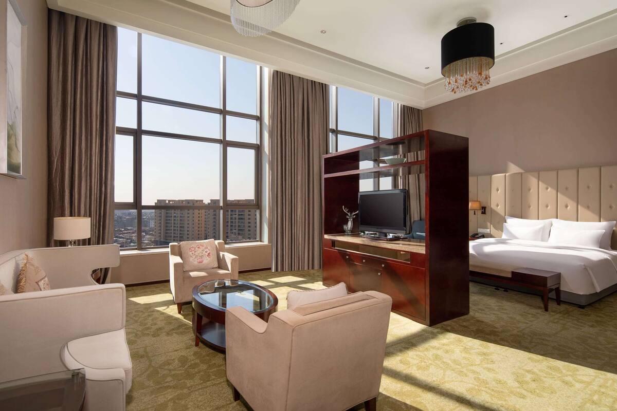 A hotel room with a large window, a bed, a desk with a TV, a sofa, and a coffee table.