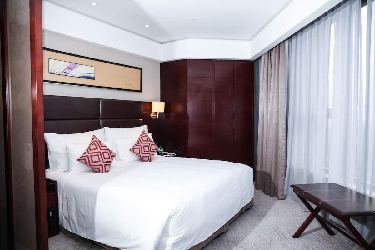 A hotel room with a large bed, a bedside table with a lamp, a wooden headboard, a framed picture on the wall, and a view of the outside through sheer curtains.