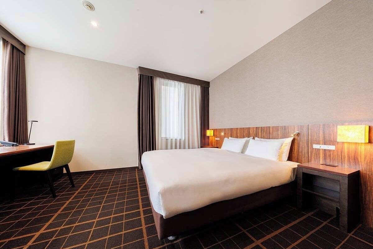 Een hotelkamer met een bed, bureau, stoel, nachtkastje en een raam met gordijnen.