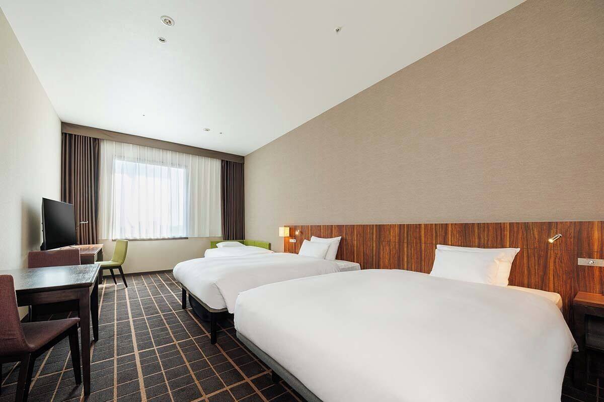 Een hotelkamer met drie bedden, een bureau, een stoel, een televisie en een raam met gordijnen.