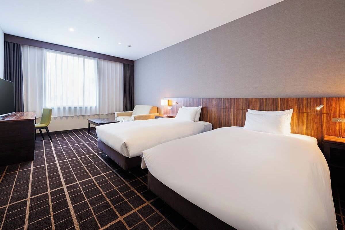 Een hotelkamer met twee bedden, een bureau, een stoel, een televisie en een groot raam met gordijnen.