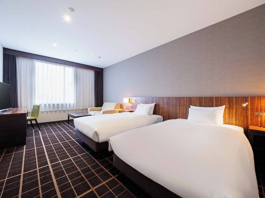 A hotel room with two beds, a desk, a chair, a TV, and a large window with curtains.