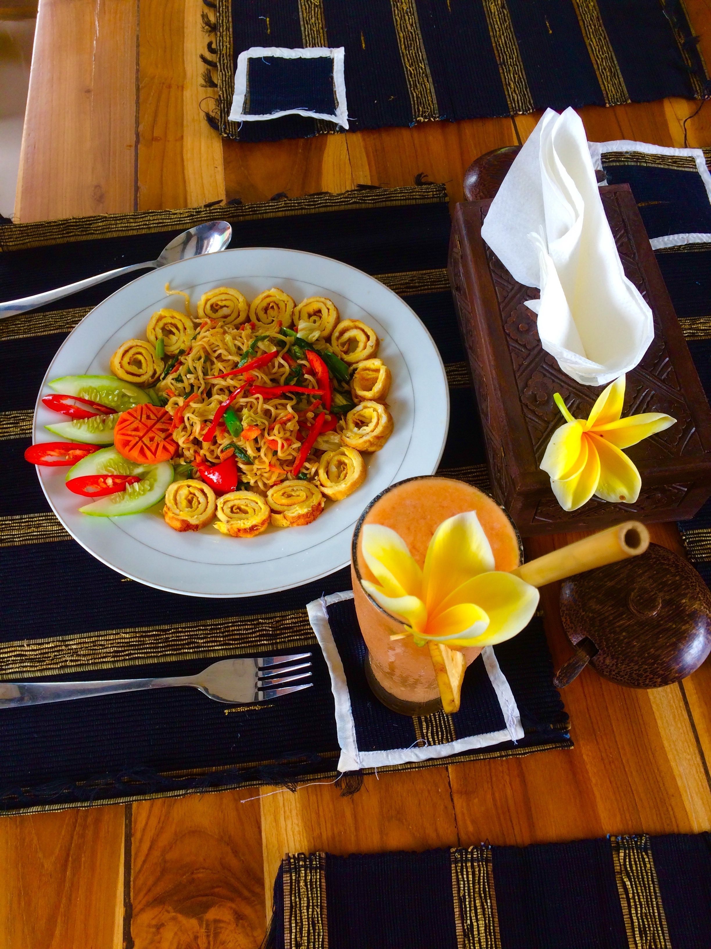 A table set with a plate of noodles, vegetables, and a glass of drink with a flower.