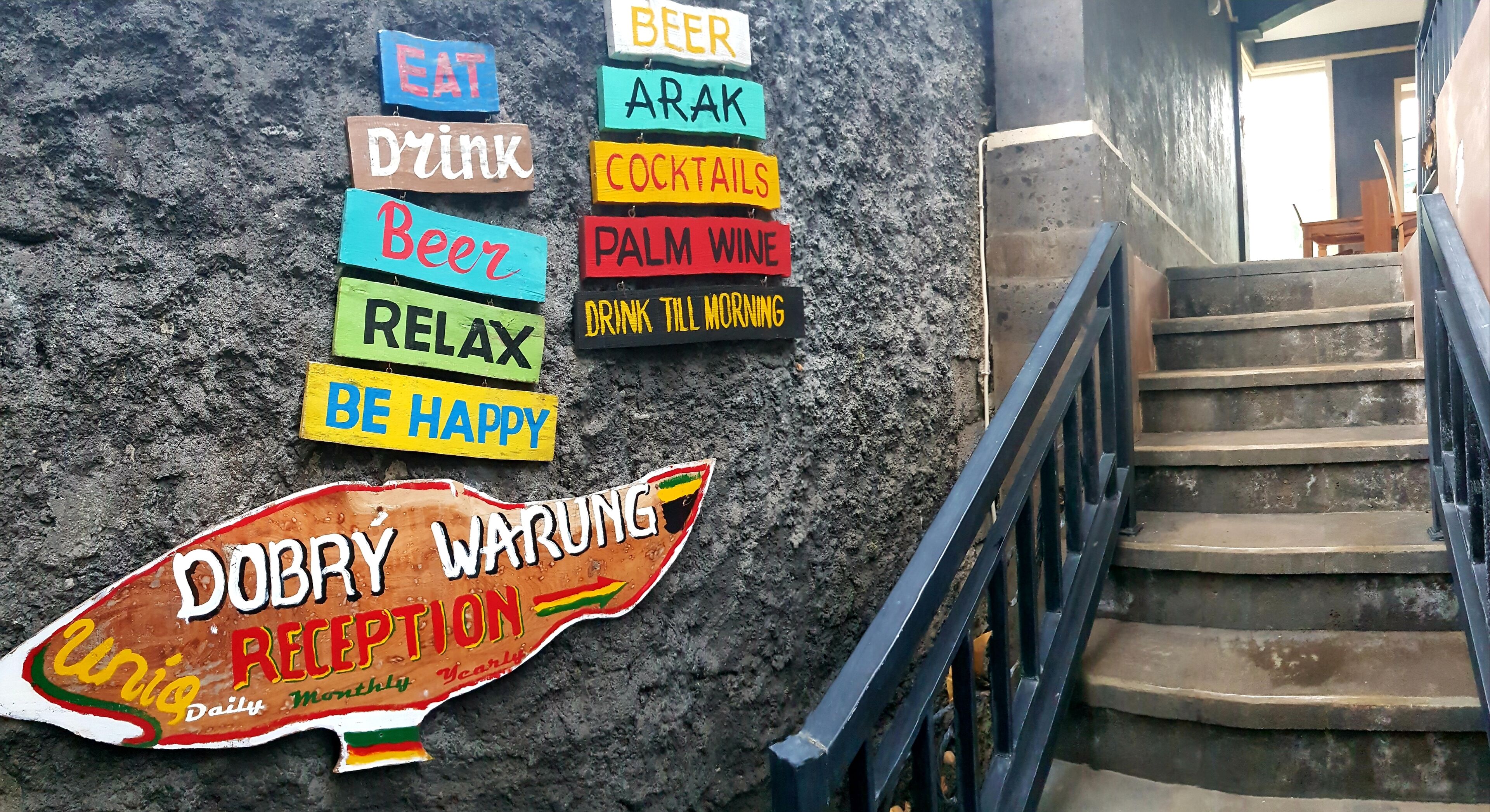 Un mur orné d’enseignes colorées faisant la publicité de diverses boissons et une pancarte indiquant l’entrée du « Dobrý Warung ».