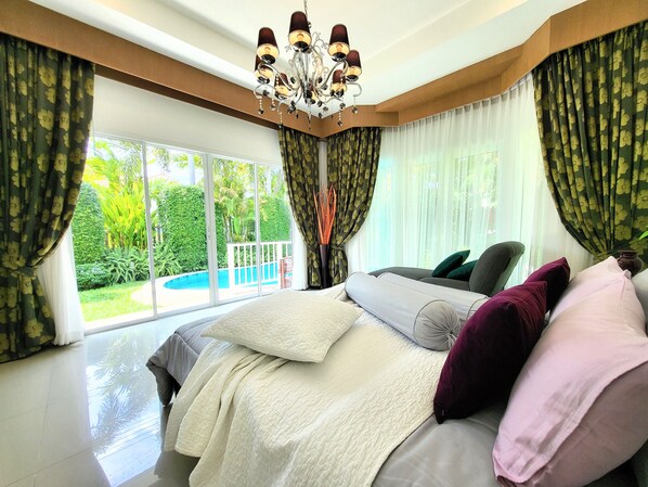 Quarto com uma cama grande, um candelabro e vista para uma piscina e vegetação.