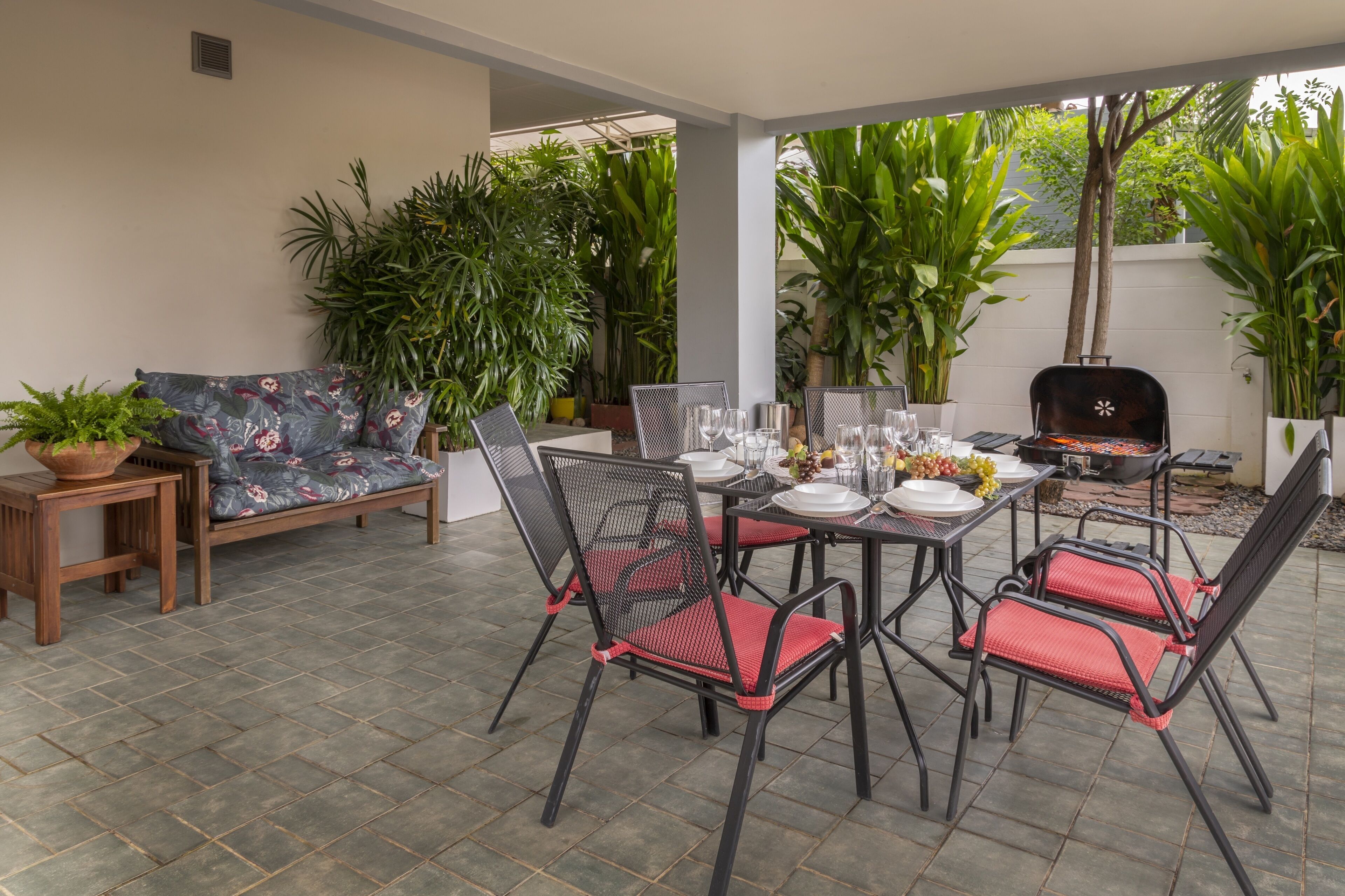 An outdoor patio with a dining table set for four, surrounded by chairs with red cushions. There is a wooden bench with a floral cushion, and potted plants are placed around the area.