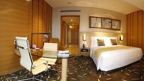 A hotel room with a glass desk, a white chair, a bed with a white and gold bedspread, and a wall-mounted artwork.