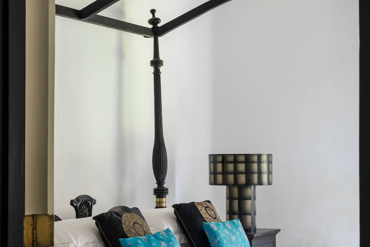 Uma cama de dossel com estrutura preta e roupa de cama branca, uma mesinha de cabeceira com abajur, e um luminário fixado na parede.