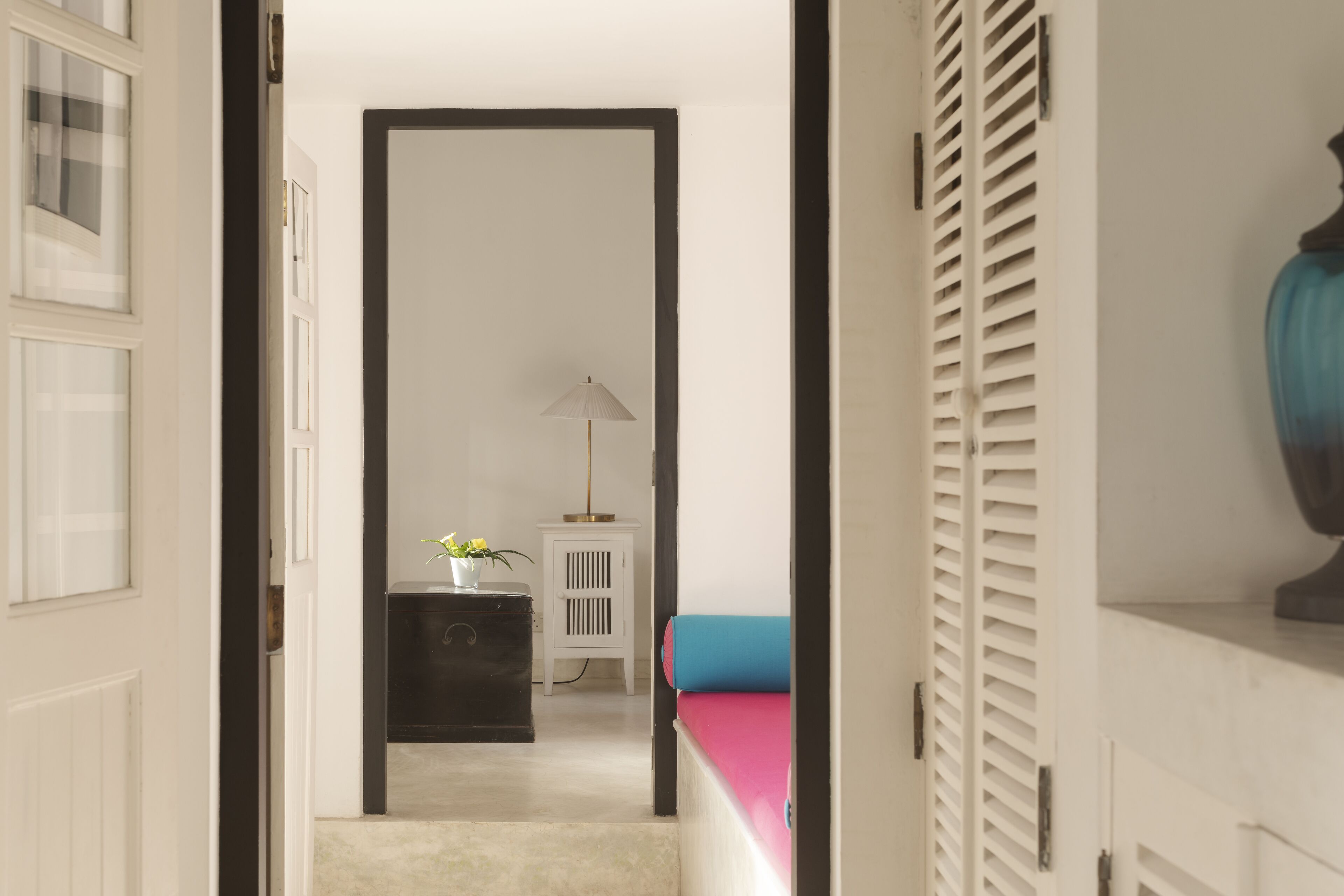Un pasillo con una puerta blanca, un armario negro, una mesita blanca pequeña y una cama azul.