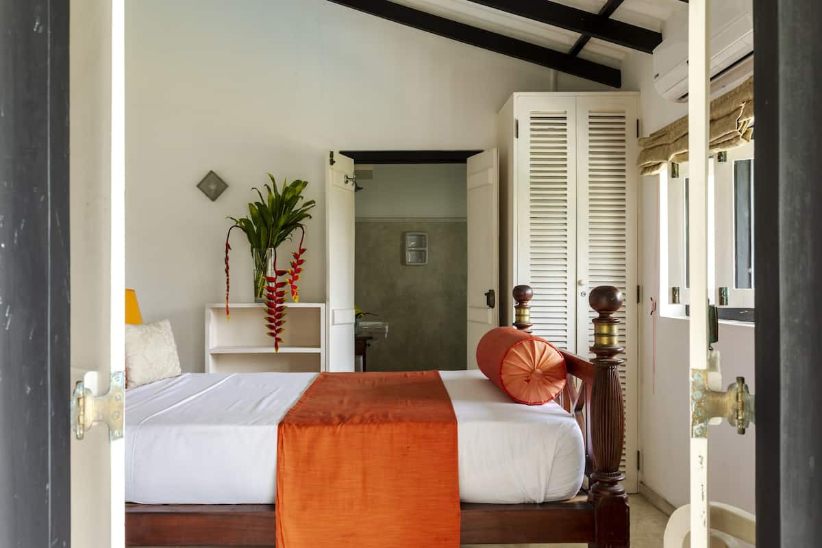 Quarto de hotel com cama, mesa de cabeceira com planta, janela com persianas e porta.