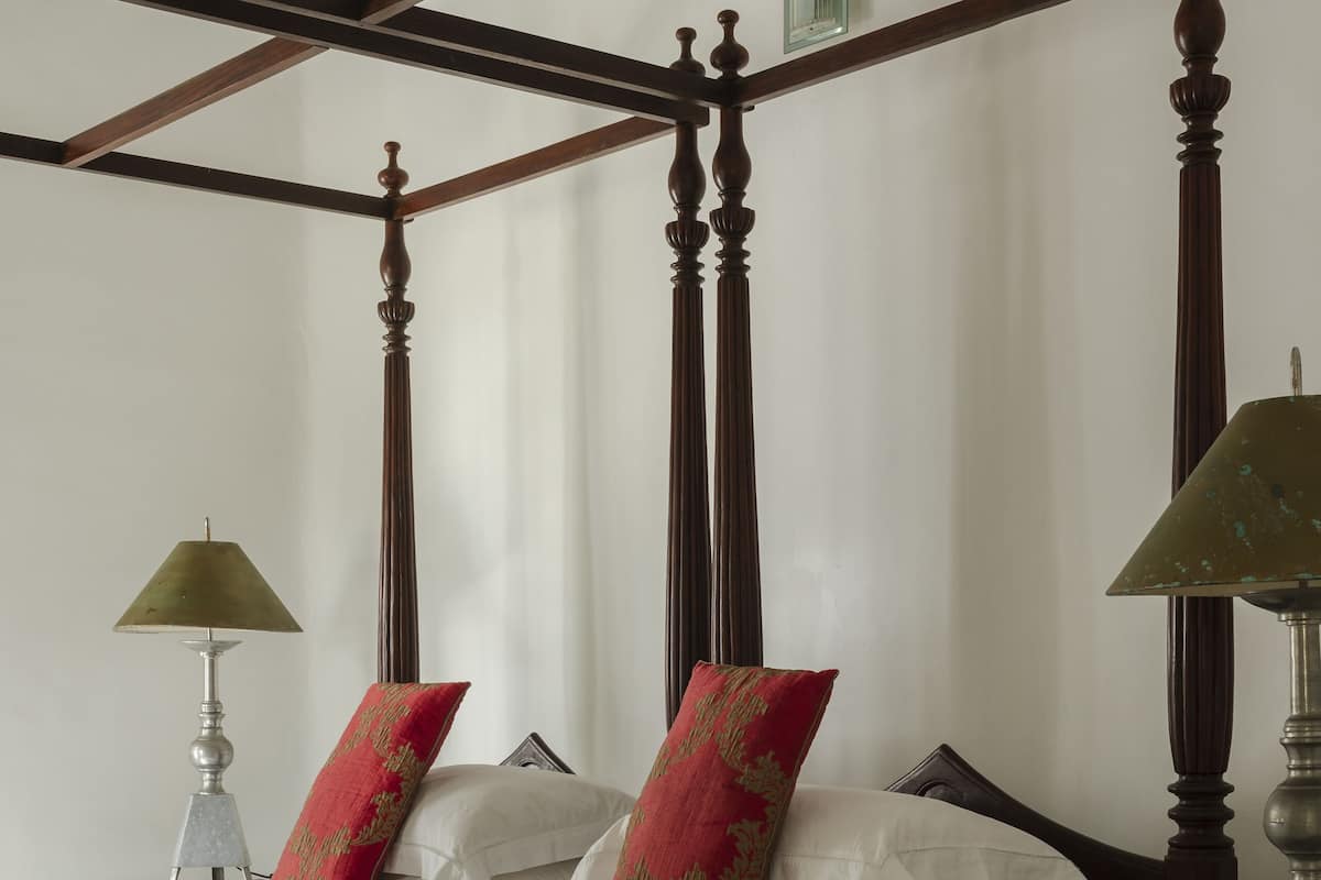 Uma cama com dossel, roupa de cama branca e almofadas vermelhas e douradas, um criado-mudo de madeira com abajur e um ventilador de teto.